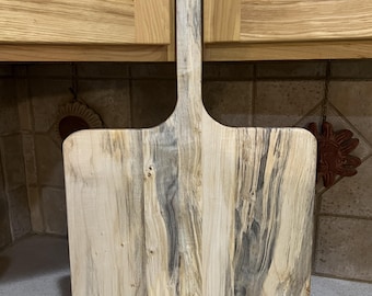 May include: A rectangular wooden serving board with a long, narrow handle featuring a circular hole at the top. The board displays a natural wood grain pattern with prominent streaks of light cream, tan, and dark gray mineral staining throughout.