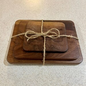 May include: A set of three wooden serving boards of varying sizes, tied together with twine. The boards are rectangular with rounded corners and a rich, dark brown color, suggesting a natural wood grain. The twine is tied in a decorative bow.