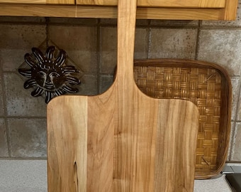 May include: A wooden pizza peel with a long handle. The peel is made of light wood with a natural grain pattern. The handle is round and has a hole at the top for hanging.