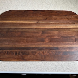 May include: Rectangular wooden cutting board with rounded corners. The board is made of dark brown wood with visible grain and is constructed from multiple wood pieces. The surface is smooth and appears to be finished.