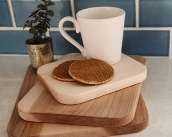 May include: Three wooden cutting boards stacked on top of each other. The top board is light brown and has a white ceramic mug and two round waffle cookies on it. The middle board is a darker brown and the bottom board is the lightest brown. The boards are on a white countertop.