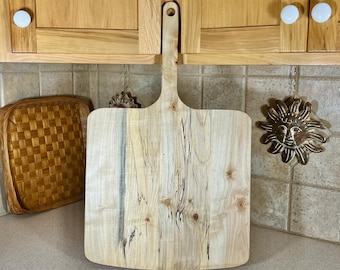 May include: A large, light-colored wooden cutting board with a handle. The board has a natural wood grain pattern with dark streaks and knots. The handle has a hole for hanging. The board is approximately 18 inches long and 12 inches wide.