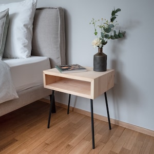 Op de afbeelding: Een lichtgekleurd houten nachtkastje met een rechthoekig ontwerp en een onderste plank. Het heeft zwarte metalen poten en een kleine vaas met bloemen erop. Een boek ligt op het oppervlak. Het nachtkastje staat naast een bed.
