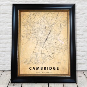Op de afbeelding: Een vintage-stijl kaartprint van Cambridge, Engeland, in een zwart frame. De kaart is beige met zwarte lijnen en tekst. De tekst onderaan luidt "CAMBRIDGE 52.1951° N - 0.1313° E".