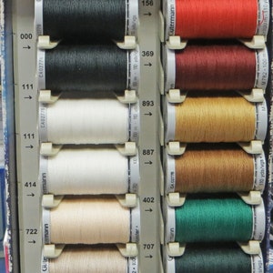 Puede incluir: Un expositor de carretes de hilo Gutermann Extra Strong en varios colores, incluyendo negro, blanco, marr&oacute;n, rojo, verde, azul y gris. Los carretes est&aacute;n etiquetados con n&uacute;meros y flechas, que indican su orden y direcci&oacute;n. El texto "Extra Strong" es visible en la parte superior del expositor.