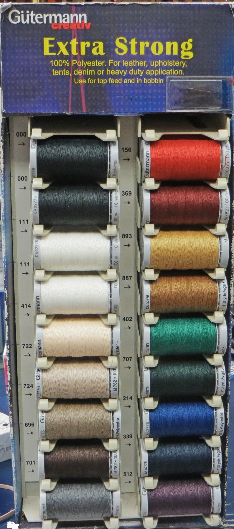 Puede incluir: Un expositor de carretes de hilo Gutermann Extra Strong en varios colores, incluyendo negro, blanco, marr&oacute;n, rojo, verde, azul y gris. Los carretes est&aacute;n etiquetados con n&uacute;meros y flechas, que indican su orden y direcci&oacute;n. El texto "Extra Strong" es visible en la parte superior del expositor.