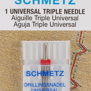 May include: A clear plastic package containing a SCHMETZ Universal Triple Needle. The package has blue and white text, including "SCHMETZ" and "1 UNIVERSAL TRIPLE NEEDLE." The needles are silver with a red plastic base. The package also includes the text "130/705 H DRI 3,0/80."