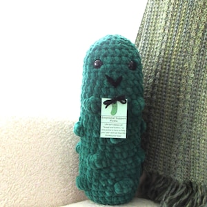 Op de afbeelding: Een donkergroene gehaakte augurk knuffel met twee zwarte plastic ogen en een kleine zwart geborduurde mond. Een rechthoekig label is met een zwart touwtje om de nek gebonden, voorzien van een augurkillustratie en de tekst Emotional Support Pickle.