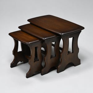 May include: A set of three nesting tables crafted from dark brown wood. Each table features a rectangular top with rounded corners and four legs with a cutout design. The tables are arranged in a descending size order, with the largest table positioned at the back and the smallest at the front, showcasing a consistent wood grain finish throughout the set.