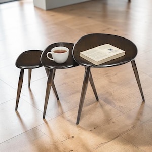 May include: A trio of dark brown nesting tables with oval tops and angled legs. One table holds a white teacup with tea, and another has a book. The tables are on a light brown wooden floor.