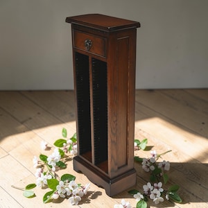 May include: A tall, dark brown wooden CD storage cabinet with a small drawer and decorative metal pull. The cabinet has multiple vertical slots for CD storage. The cabinet is on a wooden floor with white flowers.