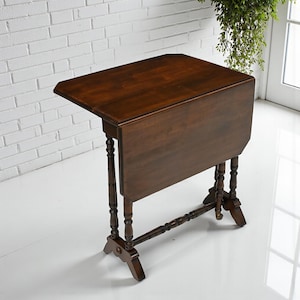 May include: A dark brown, wooden drop-leaf table with a rectangular top and turned legs. The table is in the open position, with one leaf extended. The legs are connected by a stretcher. The table is a classic piece of furniture.