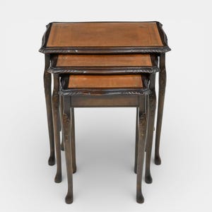 May include: A set of three antique nesting tables. Each table features a rectangular, brown leather top with a dark wood frame. The legs are carved with a floral design. The tables are nested together, with the smallest table fitting under the middle table, and the middle table fitting under the largest.