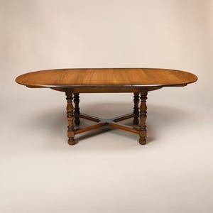 May include: An oval wooden dining table with a smooth, light brown surface and a visible grain pattern. The table is supported by four turned, spindle-style legs connected by a central X-shaped stretcher base, providing a stable foundation.