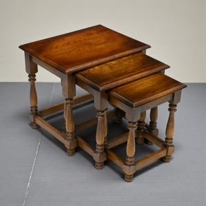 May include: A set of three wooden nesting tables in shades of brown. Each table has a rectangular top and turned legs. The tables are designed to be stored together, with the smaller tables fitting under the larger one. The tables are made of wood.