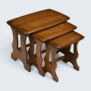 May include: A set of three nested tables crafted from medium-brown wood with a visible grain pattern. Each table features a rectangular top with rounded corners and four legs connected by a horizontal support bar, creating a tiered arrangement.