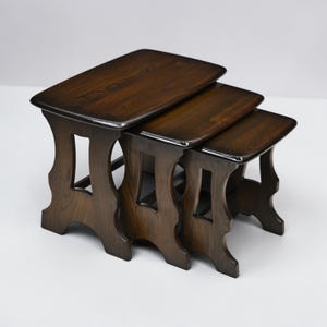 May include: A set of three nesting tables crafted from dark brown wood with a polished finish. Each table features a rectangular top with rounded corners and four legs connected by a central vertical cutout design, allowing them to stack together.