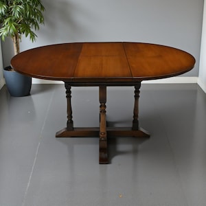 May include: Oval wooden dining table with a dark brown finish. The table has a pedestal base with four legs and a central support. The tabletop is made of three sections. A potted plant is visible in the background.