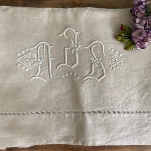 May include: A beige linen sheet with white embroidered initials, possibly a monogram. The initials are stylized with floral accents. A row of small cutouts runs along the bottom edge. A cluster of purple flowers rests in the upper right corner.