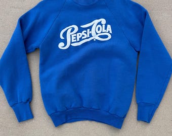 May include: A royal blue sweatshirt featuring the Pepsi-Cola logo in white script. The crew neck sweatshirt has long sleeves with ribbed cuffs and hem. The vintage-style top is made of a soft material. A classic piece of casual wear.