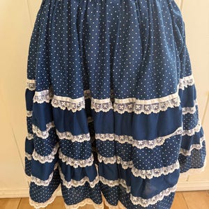 May include: A dark blue skirt featuring a white polka dot pattern, an elasticized waistband, and multiple horizontal tiers. Each tier is accented with a white satin ribbon and a delicate white lace trim along the bottom edge of the fabric.