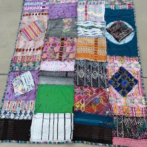 May include: A rectangular patchwork quilt constructed from various fabric squares featuring diverse patterns, textures, and colors including green, purple, teal, black, and white. The quilt displays intricate woven designs, floral motifs, and geometric shapes stitched together in a grid-like arrangement with visible seams and frayed edges.