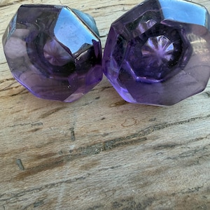 May include: Two octagonal door knobs made of translucent purple glass. Each knob features a faceted surface with a star-shaped pattern at the center, reflecting light through the deep violet material to create a clear, geometric appearance.