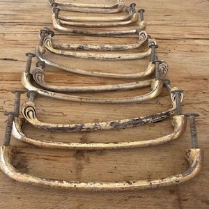 May include: A collection of antique brass drawer pulls with a curved design. The pulls have a distressed, aged appearance with visible wear and tear, and are mounted with screws. The hardware is arranged on a wooden surface.