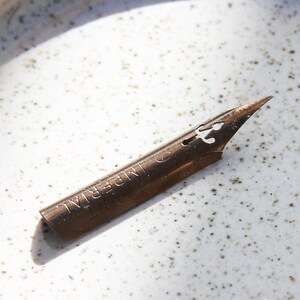 Könnte beinhalten: Eine antike, bronzefarbene Feder mit dem Wort "IMPERIAL" auf der Seite. Die Feder hat eine spitze Spitze und einen dekorativen Ausschnitt in der Nähe der Oberseite. Sie liegt auf einer gesprenkelten, cremefarbenen Oberfläche.