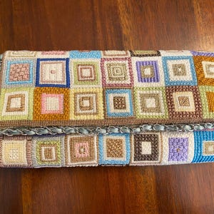 Puede incluir: Un bolso de mano rectangular y colorido con un diseño de patchwork. El bolso presenta un patrón repetitivo de cuadrados en varios colores, incluyendo azul, rosa, verde y marrón. Los bordes están adornados con una cadena plateada. Un accesorio elegante.
