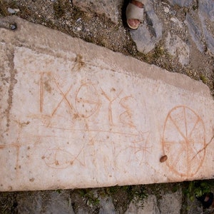 May include: A weathered stone slab with faded orange markings. The markings include geometric shapes, lines, and what appear to be ancient symbols or letters. The stone is light in color and appears to be part of a larger structure.