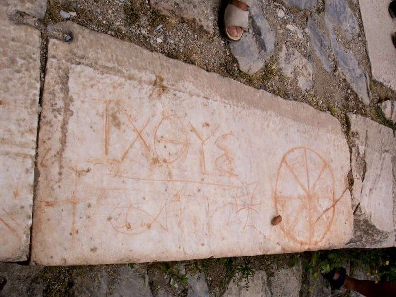 May include: A weathered stone slab with faded orange markings. The markings include geometric shapes, lines, and what appear to be ancient symbols or letters. The stone is light in color and appears to be part of a larger structure.