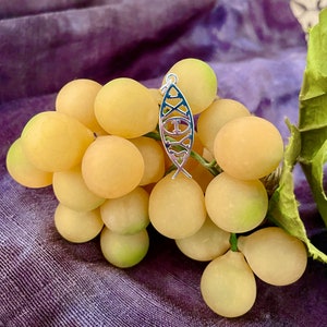 May include: A silver-colored pendant in the shape of a fish, with a blue and white design, hangs over a bunch of yellow grapes. The pendant has a loop at the top for a chain. The grapes are on a purple background.
