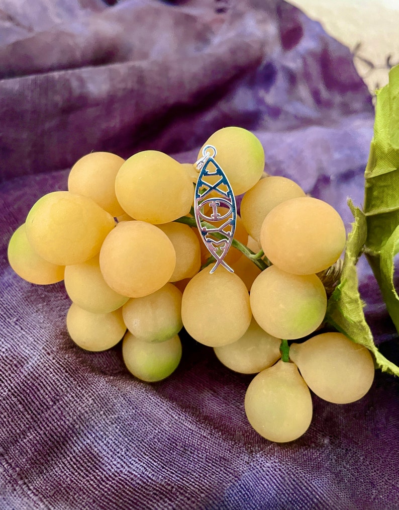 May include: A silver-colored pendant in the shape of a fish, with a blue and white design, hangs over a bunch of yellow grapes. The pendant has a loop at the top for a chain. The grapes are on a purple background.