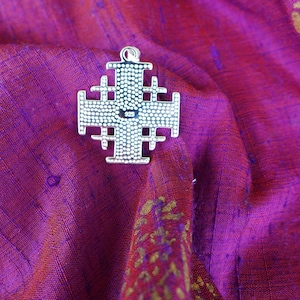 May include: A silver Jerusalem cross pendant with a detailed design. The cross is on a purple fabric background.