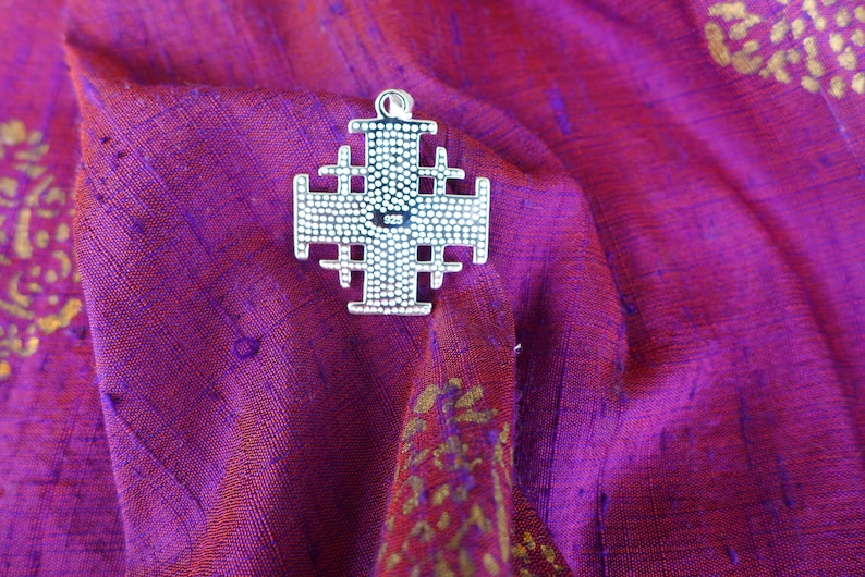 May include: A silver Jerusalem cross pendant with a detailed design. The cross is on a purple fabric background.