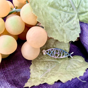 May include: A silver-colored pendant in the shape of a fish, with the Greek letters 'IX&Theta;Y&Sigma;' (Ichthys) inside. The pendant rests on a green leaf and a purple fabric, with a bunch of light yellow grapes in the background.