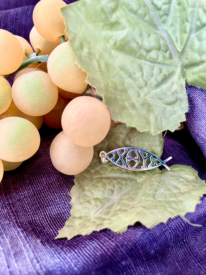 May include: A silver-colored pendant in the shape of a fish, with the Greek letters 'IX&Theta;Y&Sigma;' (Ichthys) inside. The pendant rests on a green leaf and a purple fabric, with a bunch of light yellow grapes in the background.
