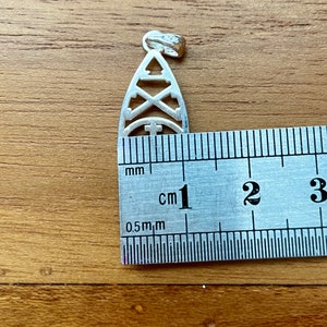 May include: A silver pendant with a cut-out design featuring geometric shapes, including a triangle, crossed arrows, and a cross within a semi-circle. The pendant is approximately 1 inch in length and has a small loop at the top for a chain.