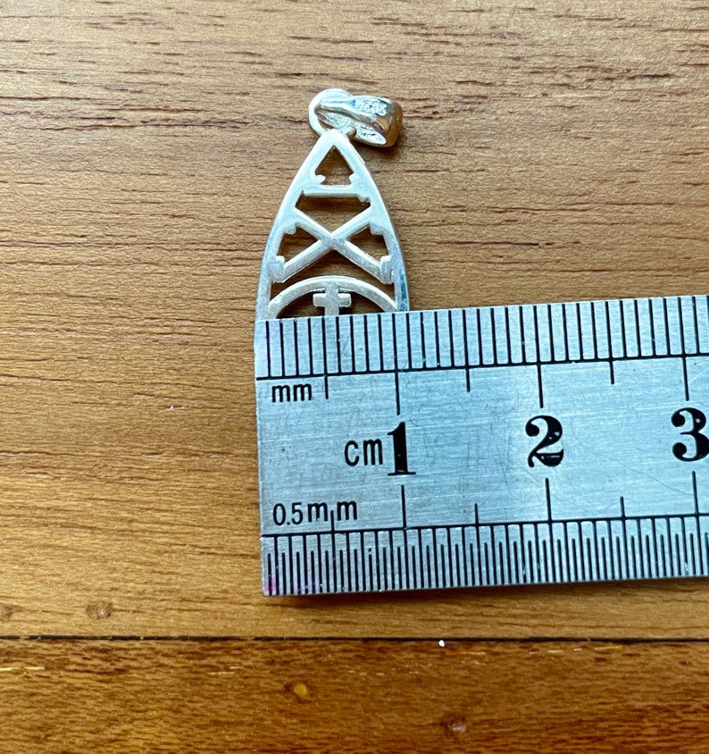 May include: A silver pendant with a cut-out design featuring geometric shapes, including a triangle, crossed arrows, and a cross within a semi-circle. The pendant is approximately 1 inch in length and has a small loop at the top for a chain.