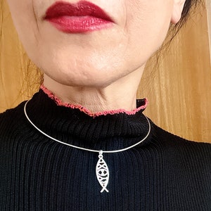 May include: A silver-colored pendant necklace featuring a stylized fish design. The pendant hangs from a thin, silver-colored choker. The person in the image is wearing a black sweater with a pink trim.