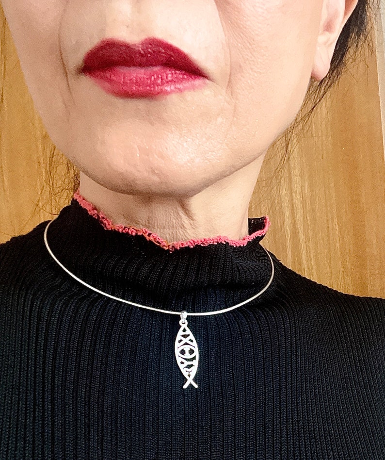 May include: A silver-colored pendant necklace featuring a stylized fish design. The pendant hangs from a thin, silver-colored choker. The person in the image is wearing a black sweater with a pink trim.