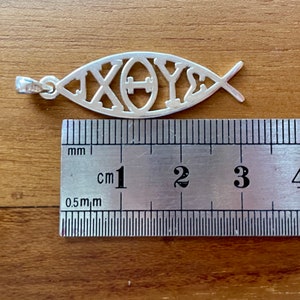 May include: A silver pendant in the shape of a fish, with the Greek letters Chi, Theta, and Upsilon, and Sigma, measuring approximately 1.75 inches in length. The pendant is displayed on a wooden surface next to a ruler.