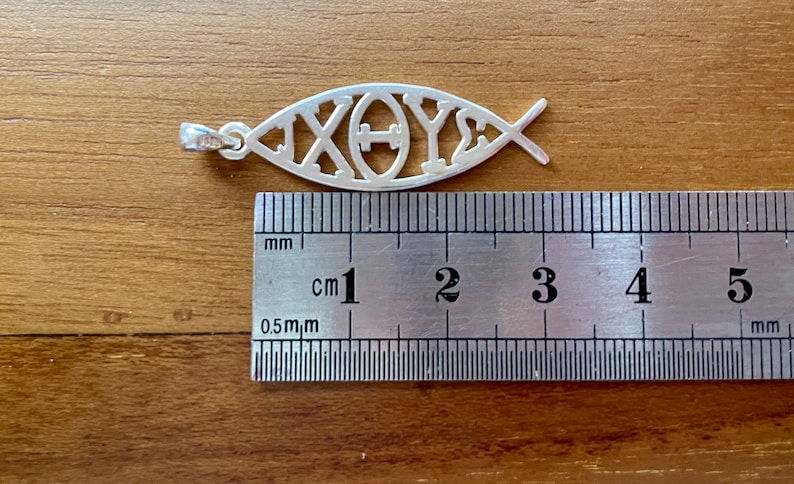 May include: A silver pendant in the shape of a fish, with the Greek letters Chi, Theta, and Upsilon, and Sigma, measuring approximately 1.75 inches in length. The pendant is displayed on a wooden surface next to a ruler.