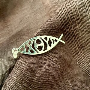 May include: A silver pendant in the shape of a fish, with the Greek letters Chi, Rho, Theta, and Upsilon, resting on a textured brown fabric. The pendant has a small loop for a chain.