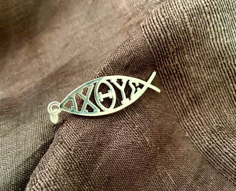 May include: A silver pendant in the shape of a fish, with the Greek letters Chi, Rho, Theta, and Upsilon, resting on a textured brown fabric. The pendant has a small loop for a chain.