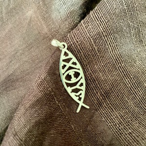May include: A silver pendant in the shape of a fish, with intricate cut-out designs. The pendant is suspended on a small silver loop. The background is a dark brown, textured fabric.