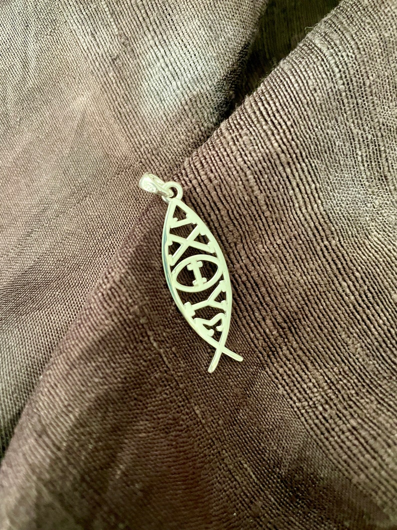 May include: A silver pendant in the shape of a fish, with intricate cut-out designs. The pendant is suspended on a small silver loop. The background is a dark brown, textured fabric.