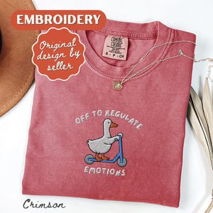 May include: Crimson-colored t-shirt with an embroidered design of a duck riding a blue scooter, with the text "OFF TO REGULATE EMOTIONS". The shirt has a crew neck and short sleeves. A gold necklace is draped across the shirt.