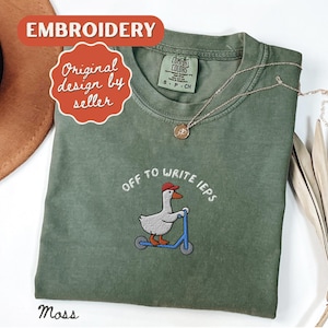 Könnte beinhalten: Moosgrünes T-Shirt mit gesticktem Design. Das Design zeigt eine Gans mit roter Kappe auf einem blauen Roller, mit dem Text "off to write IEPs". Eine goldene Halskette liegt auf dem Shirt.